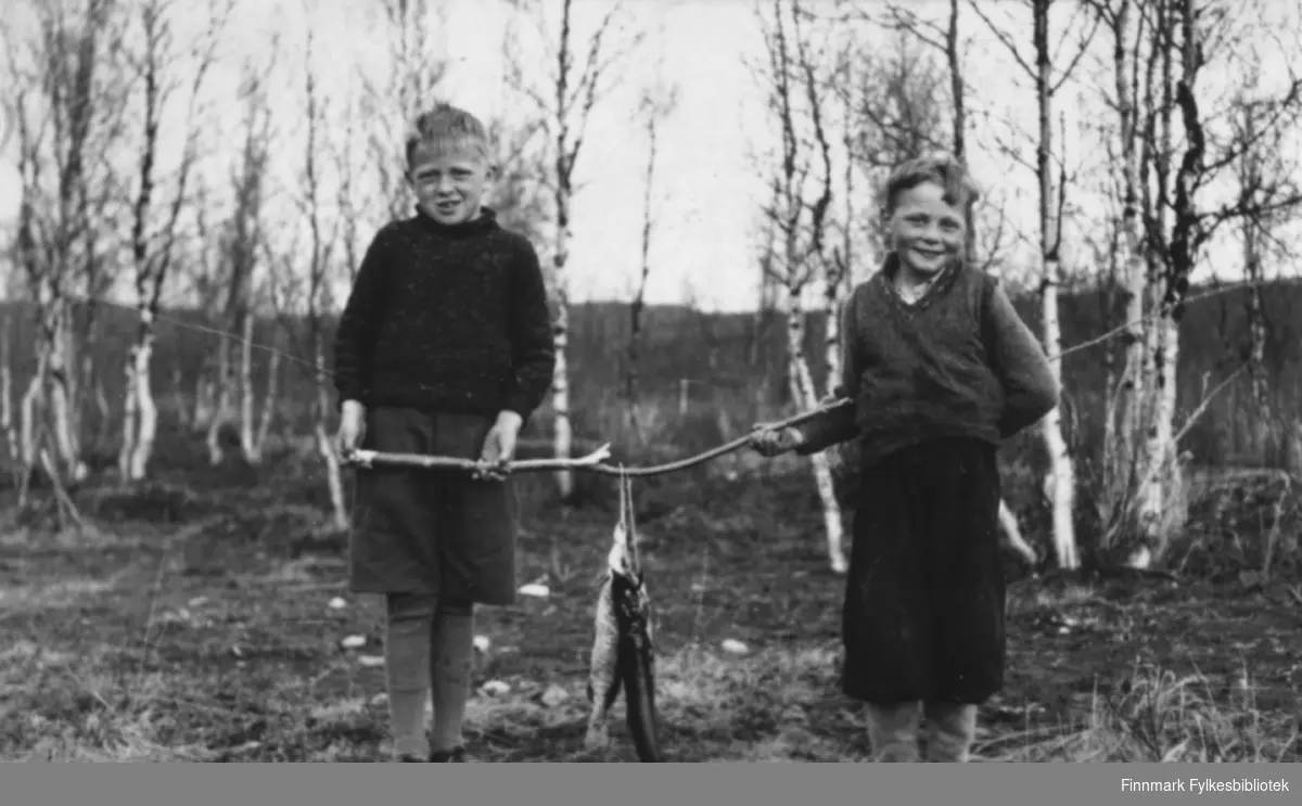 To gutter har vært på fisketur i Øvre Neiden. Gutten venstre heter antakelig Arne Valen, til høyre ser vi Kåre Sivertsen. De bærer fangsten mellom seg på en lang kjepp