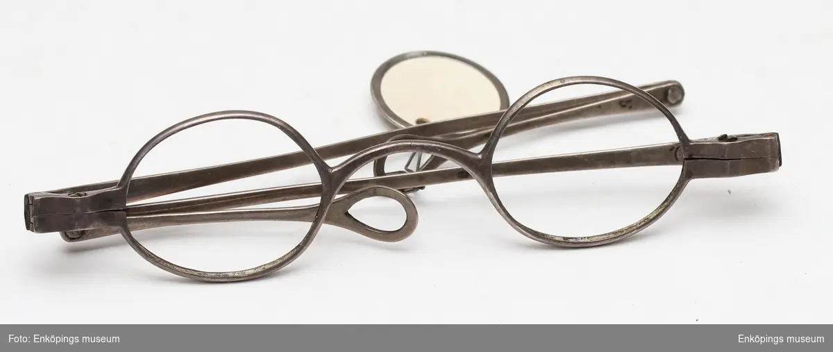 Ett par glasögon med hopfällbara skalmar av silver. Skalmarna slutar i en ögla. Saknar glas. Enkelt, något svängt nässtycke. Troligen tillverkade under 1800-talet.