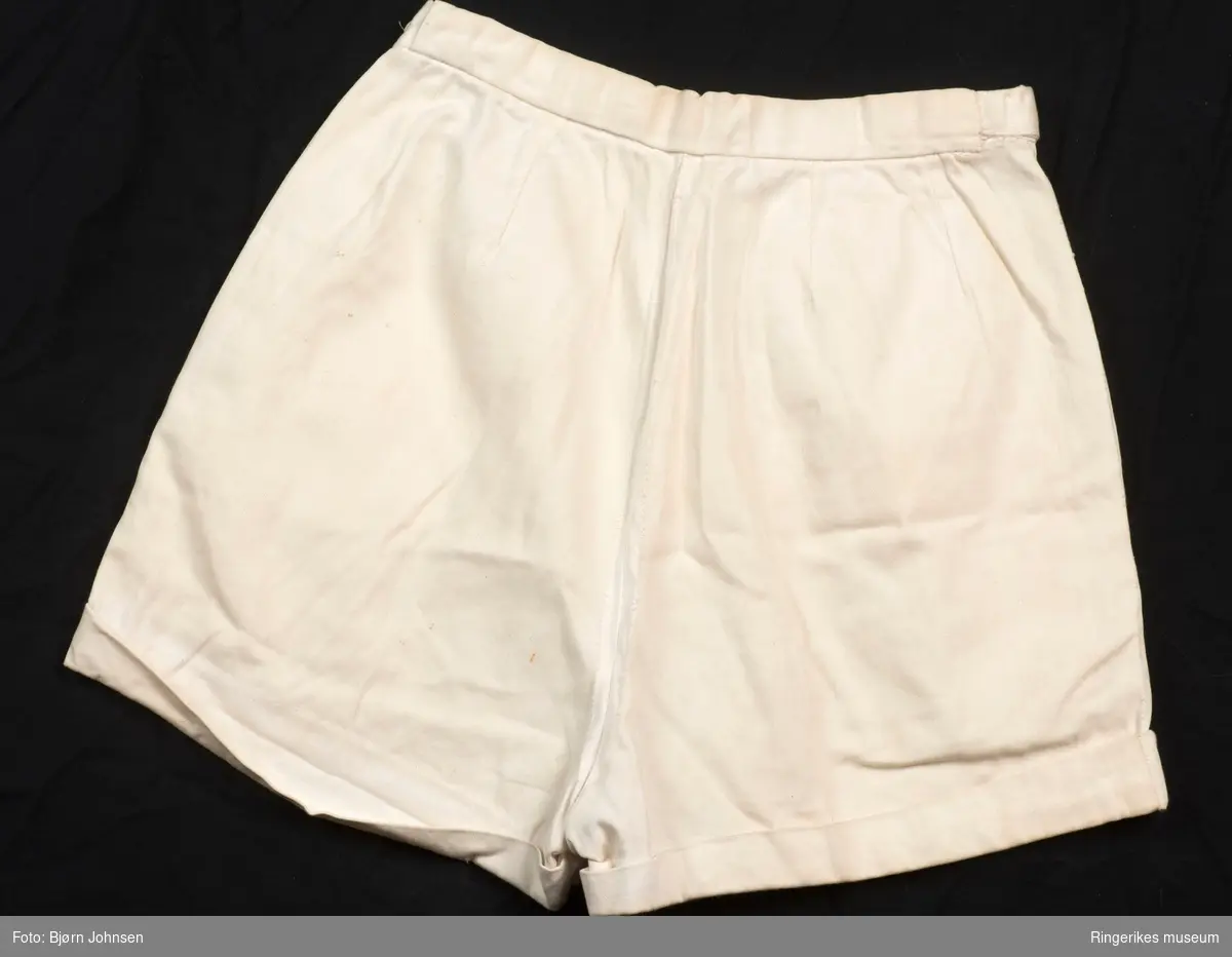 Kort bukse med oppbrett, to skrådde og dype stikklommer, glidelås på venstre side, smal linning med knapp/ knapphull og regulering på siden.