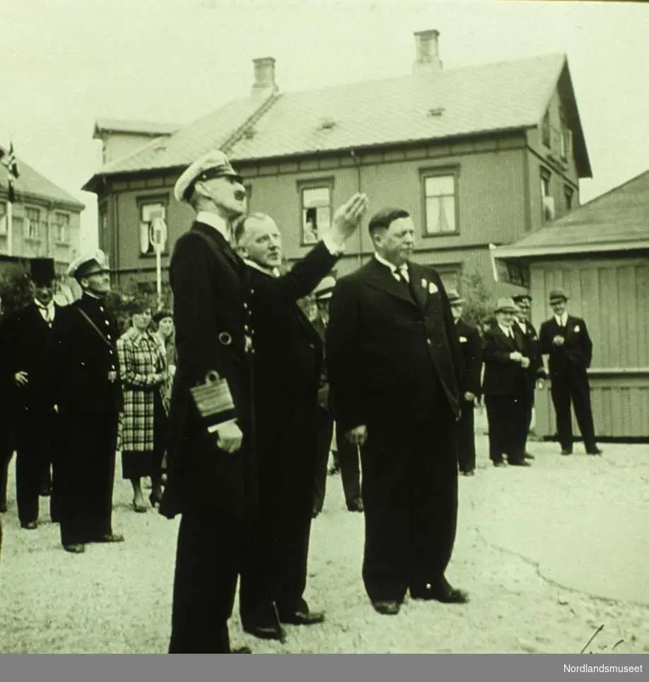 Kong Haakon på varemessa i 1937, i middelskolebakgården. Kongen betrakter et norgeskart i relieff i full lengde, som var oppslått på veggen til middelskolen. Bokbinder Falcks hus bak.