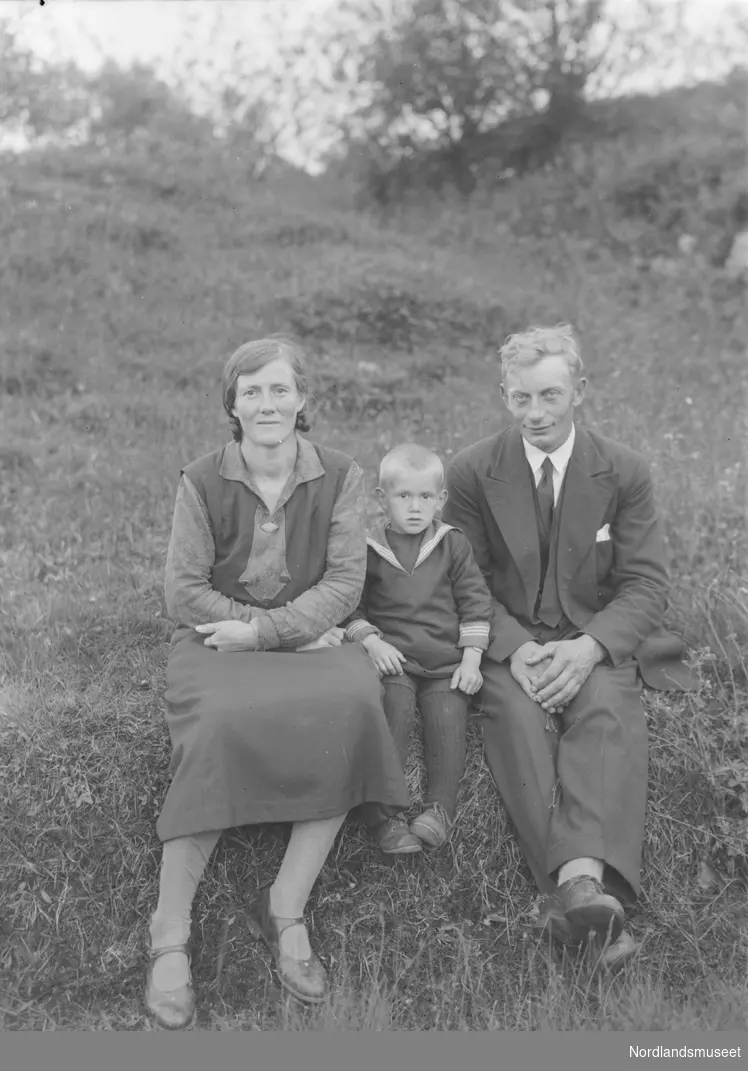 Portrett. En mann i dress og en kvinne i kjole sitter med en liten gutt mellom seg i gresset.