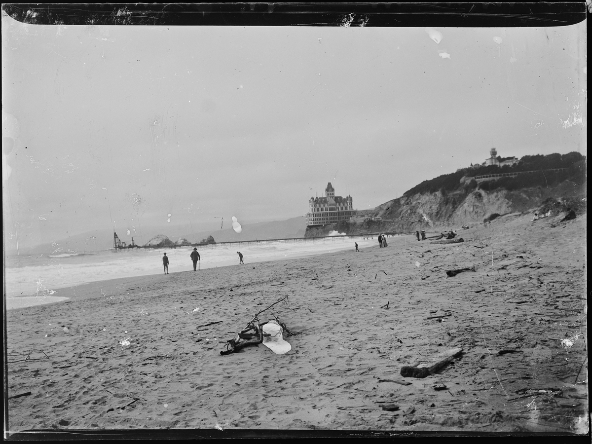 Ocean Beach, San Fransisco. Old Cliff House Restaurant på klippen i bakgrunnen, mennesker går på stranden. En lang pir går utover i havet.