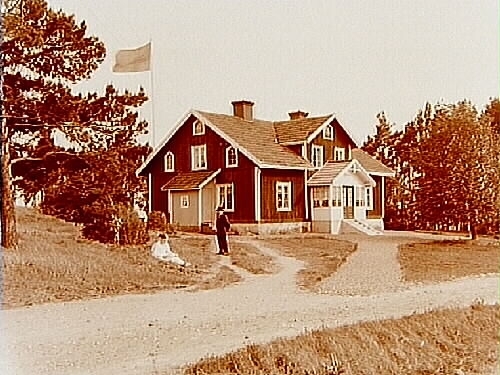 En och en halvvånings bostadshus med frontespis, glasad förstubro och förstuga.
2 personer framför huset.
Johan Karlsson