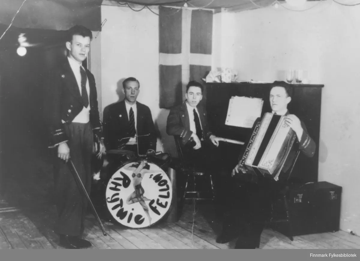 Bilde av 'Rhytmie Fellows' fra Kirkenes. Det står ingen dato på bildet. Fire menn med fiolin, trommer, piano og trekkspill. I bakgrunnen henger et flagg. Det må være dansk, svensk, eller finsk. Gruppen er kledt i dress.
