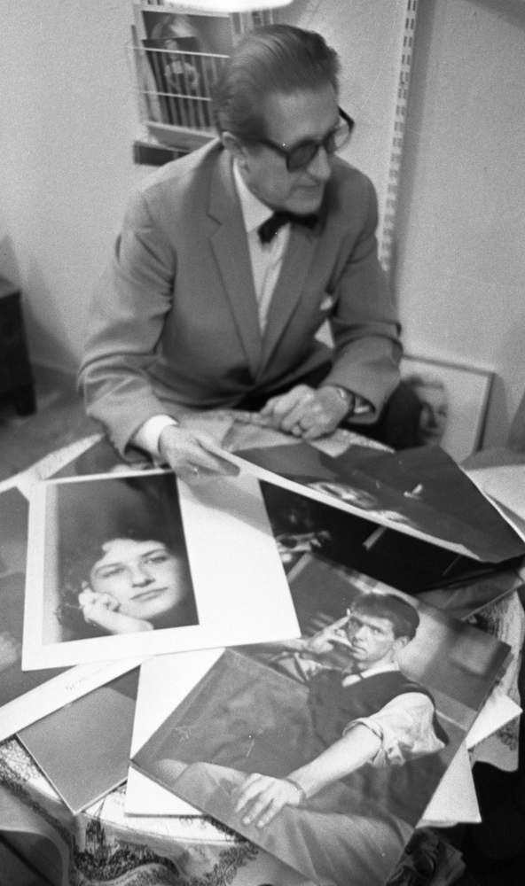 Götlin, 30 september 1965

En fotograf sitter vid ett bord och sorterar fotografier. Han är klädd i ljus kavaj samt vit skjorta med fluga till. Han bär glasögon. Bakom honom sitter en vägghylla fyllda av foton i olika fack. På golvet till höger står ett inramat fotografi.