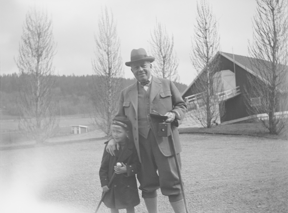 Christian Pierre Mathiesen og et barn står sammen på gårdsplassen på Linderud Gård. Mannen har lagt hånden sin på barnets skulder. Begge ser mot fortografen og smiler. I bakgrunnen sees store løvtrær uten blader og låve med låvebru.
