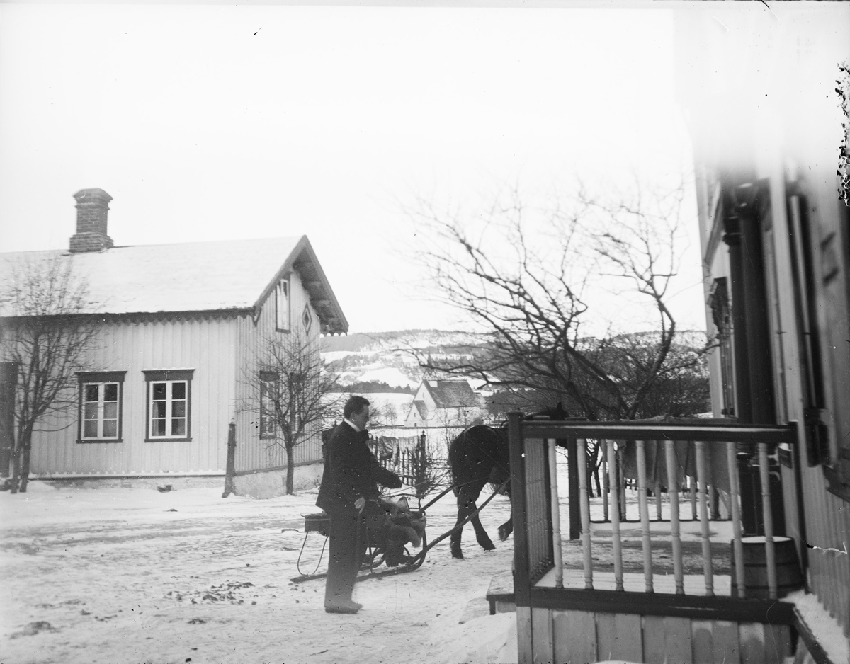 På en gårdsplass, mellom to hus, i et vinterlandskap, drar en hest en liten slede og Christian Pierre Mathiesen går fra sleden mot et av husene. Kirken i bakgrunnen er Stiklestad kirke. Bildet er tatt fra tunet på Stiklestad Nordre.