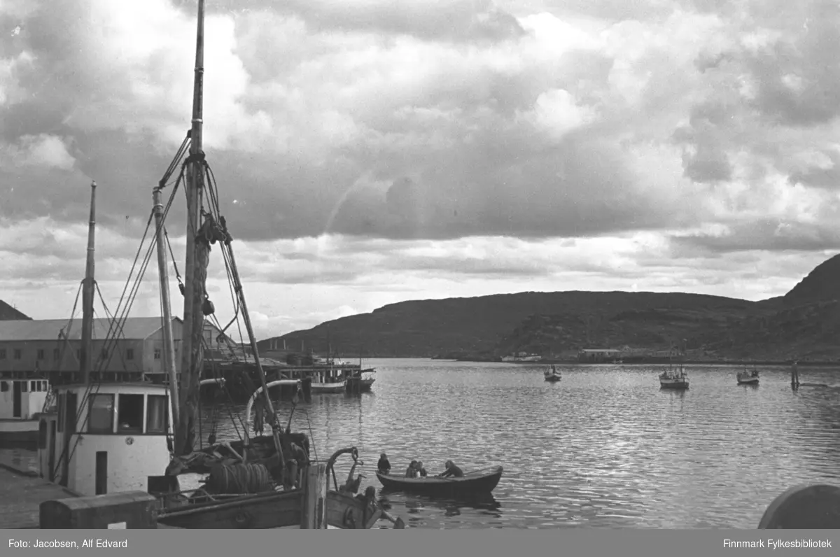 En vindstille, men overskyet sommerdag på havna i Havøysund. En båt ligger ved kaia nærmest. Båten har hvitmalt styrhus og en stor mast med bom foran og en mindre mesanmast bak. Foran på baugen står trommelen til vinsjen og litt av dreggen kan også ses. En kasse står på kaia ved siden av båten. På havna rett utenfor ligger en robåt med tre personer ombord. Robåten er av tre og har et hvitmalt bord ved rekka. En avlang brakke står på en kai til venstre på bildet. Det er Anton Olsens materialhandel som senere ble omgjort til fiskemottak. Det hvite styrhuset til en båt ses liggende ved den kaia. Et mindre fartøy ligger ved kortenden av kaia. Utpå havna ligger flere båter fortøyd og et sjømerke står langt ute på havna, helt til høyre på bildet. Et fjellparti ligger tvers over havna. Bygget som ses i strandkanten der er Haagensen-bruket.