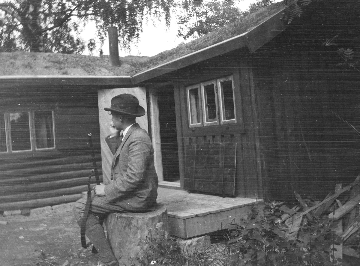 Iacob Ihlen Mathiesen sitter på en stor stubbe utenfor en tømmerhytte med torvtak. På bakken, mellom bena, står et jaktgevær.
