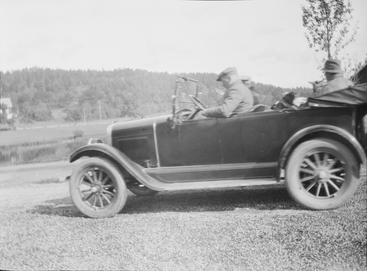 Bil fotografert på gårdsplassen til Linderud Gård. To personer sitter foran i bilen og en piperøkende person sitter i baksetet.