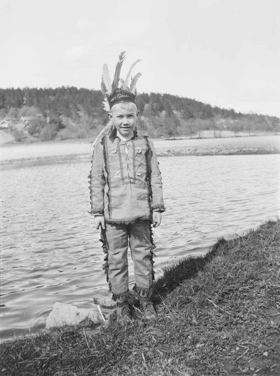 Iacob Ihlen Mathiesen står ved en gresskledd vannkant kledd i et indianerkostyme: bukse overdel og fjærpynt på hodet. I bakgrunnen sees en lav ås med gårdsbebyggelse og en landevei/gårdsvei.