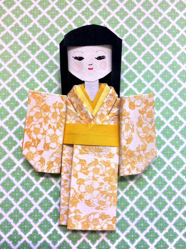 Papirdukke brettet i japanske origamiteknikk. Dukken har fått kimono i gult, blomstret papir.