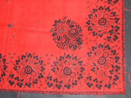 Schal av rött ylletyg med tryckt mönster i  form av svart blomsterbård runt om. Fragment av frans i rött ullgarn på sidorna. Handfållad.

Sökord: ull.