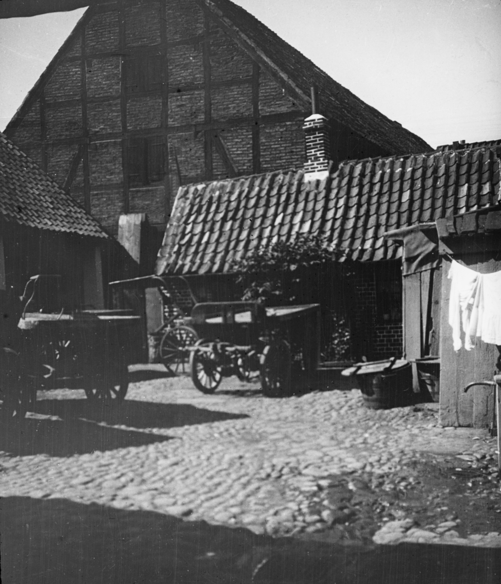 Skioptikonbild med motiv från Helsingborg. Kullerstensbeklädd bakgård med droskor och vagnar.
Bilden har förvarats i kartong märkt: Helsingborg 8. 1908
