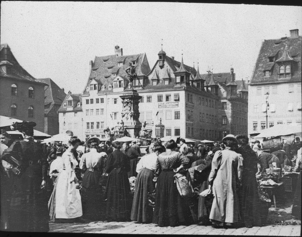 Skioptikonbild med motiv av folksamling på marknad på Hauptmarkt i Nürnberg.
Bilden har förvarats i kartong märkt: Resan 1907. Bamberg 2. Nürnberg 6. 295. Text på bild: "Hauptmkt.".