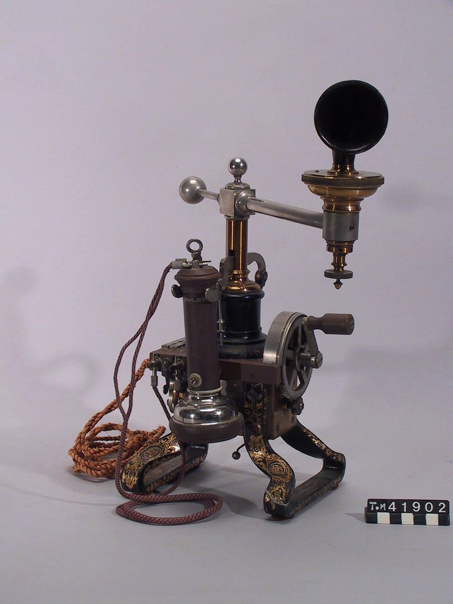 Telefonapparat, bordsmodell för lokalbatteri, med spiralmikrofon på svängbar arm. S.k. Tax. I denna telefon från 1884 utgörs de krumma benen av magneterna till ringsignalinduktorn. Den av Lars Magnus Ericsson konstruerade spiralmikrofonen sitter på en svängbar arm.