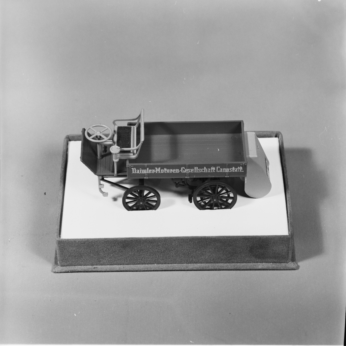 Modell av lastbil, gjuten i plast, beigefärgad med bruna hjul och svart chassi, flaklemmar med text. Hölje av plast. Lastbilens längd: 100 mm, bredd: 35 mm, höjd: 35 mm. Höljets längd: 120 mm, bredd: 85 mm, höjd: 68 mm. Märke: Daimler. Årgång: 1896.