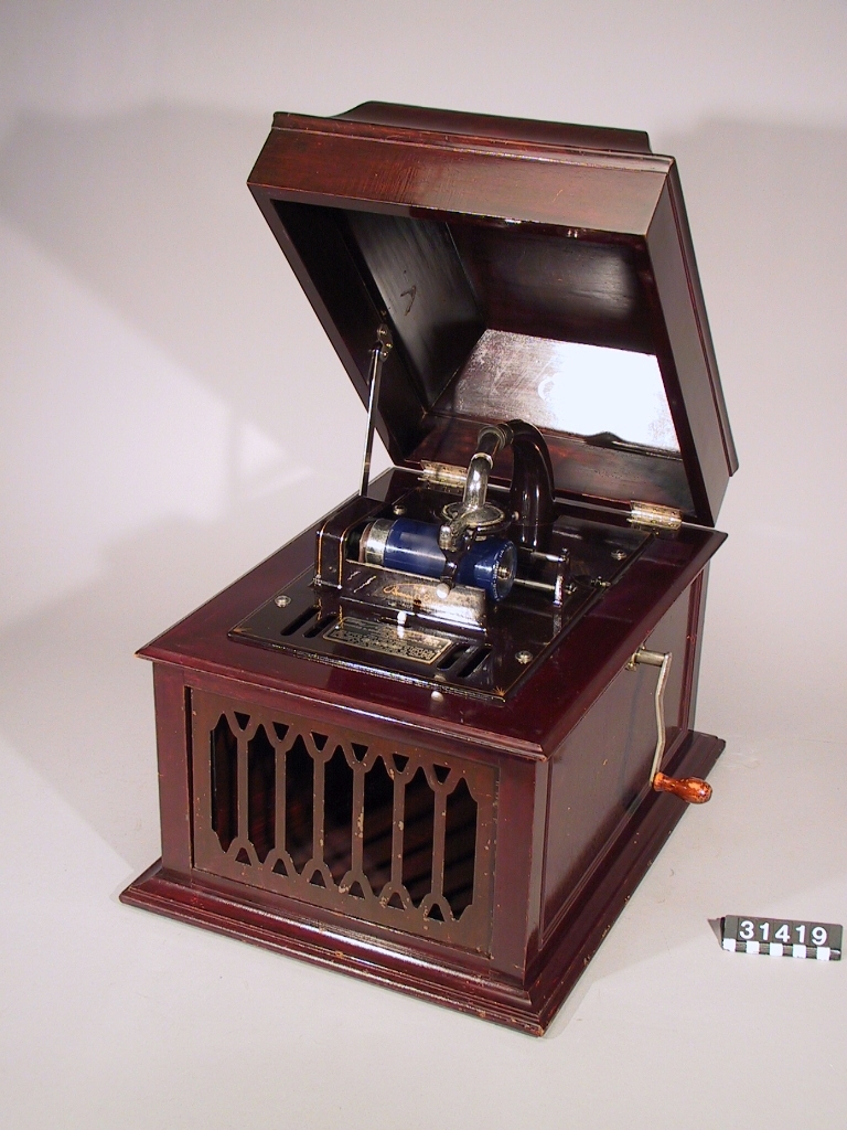 Fonograf inbyggd i låda av mahogny med uppfällbart lock, med tratt direkt från ljudarmen och utmynnande i lådans framsida. Gallret vid trattens mynning sitter löst