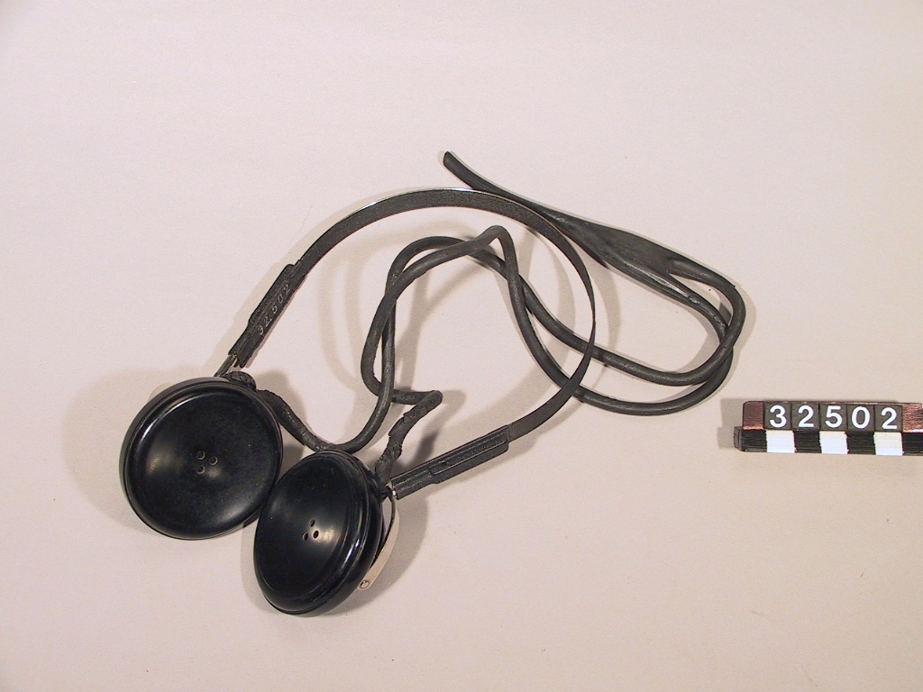 Dubbel hörtelefon av bakelit, stål-hjässfjäder och svart gummi-anslutningssladd, med ställbar hjässfjäder. Till Websters tråddiktafon.