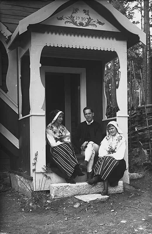 Bostadshus. Grupp tre personer i folkdräkter på trappan till huset.
Karl Hedström