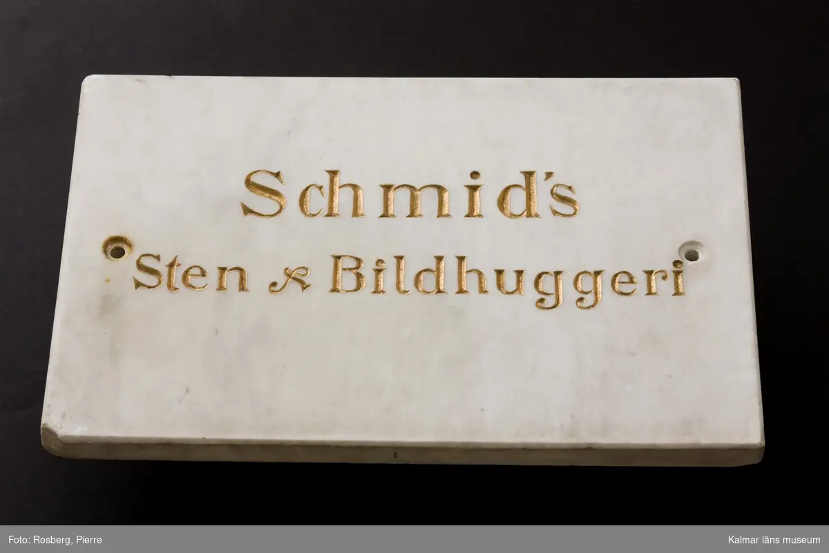 KLM 12433. Skylt, stenhuggarskylt, bildhuggarskylt. Av marmor med text i guld. Marmortavla med försänkta bokstäver i guld. Text på båda sidor, på ena sidan: J.A. Schmid, Bildhuggare en Tr. upp, på andra sidan: Schmid´s Sten & Bildhuggeri.
