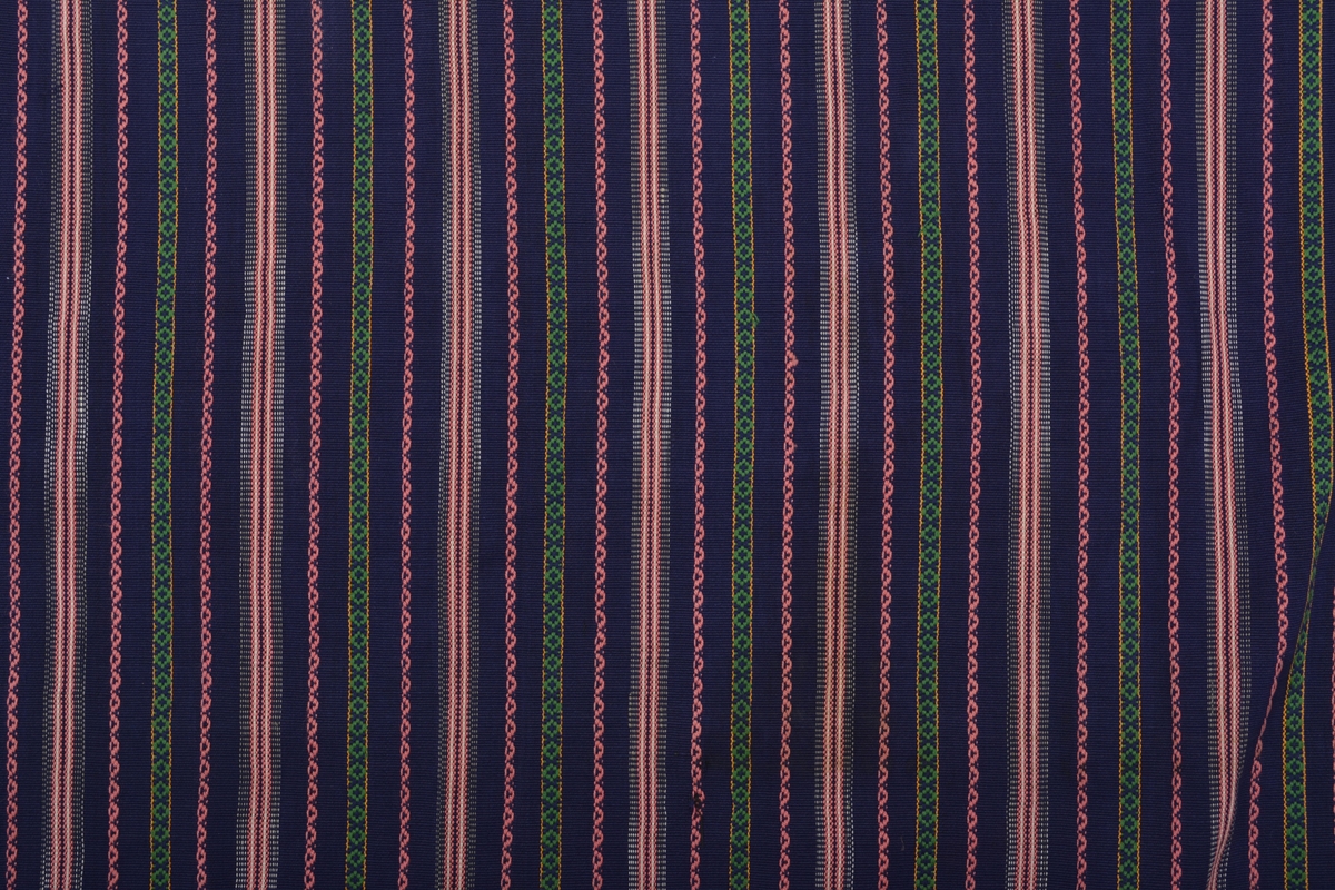 Rosengångsförkläde vävt i bomull med mörkblå botten, smala ränder i rosa/vitt samt rosengångspartier rosa/mörkblått och grönt/mörkblått/gult. Bredd nertill 895 mm, rynkat upptill vid linningen som består av ett band i samma mörkblå, rosa och vita bomullsgarn som i förklädet, dubbelvikt över linningen (10 mm syns), sedan slätt som knytband 660 mm (höger) resp 745 mm vänster. Avslutning med två flätor och tofs inräknat (40 mm). Fåll nedtill 20 mm, sidorna 4 mm. 
 
Eventuellt från Helsingborg enligt notering.
