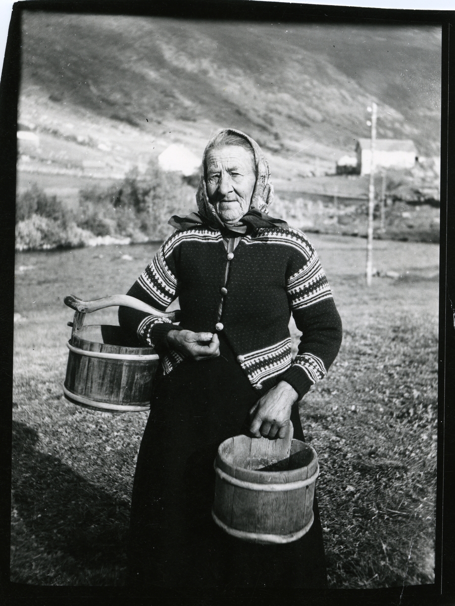 Portrett av kvinne iført skaut og lusekofte. I hendene holder hun noen trespann, såkalte mjølkeringje og kalvask fra Elvheim i Øye i Vang kommune.