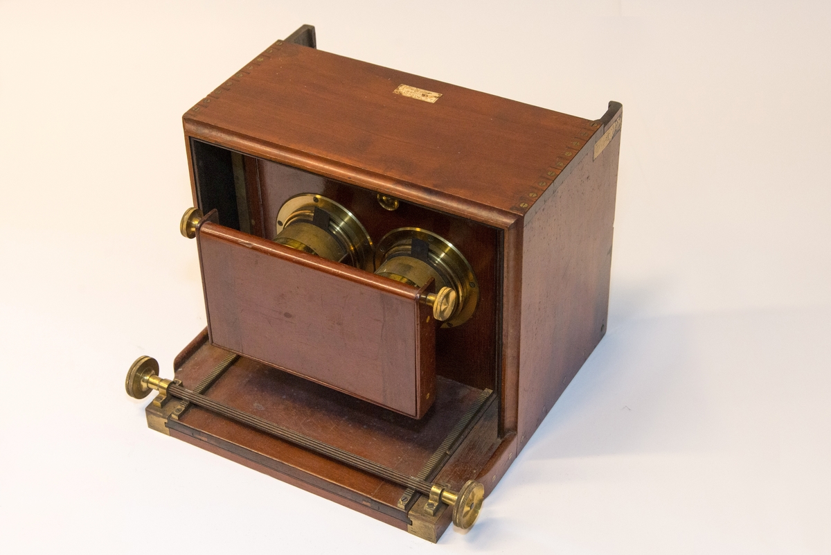 Gruppering med fotokamera i oppbevaringskasse, separat metall-lokk, stereolinse, lause delar som negativplate, mattskive, blendarsett og diverse smådelar.