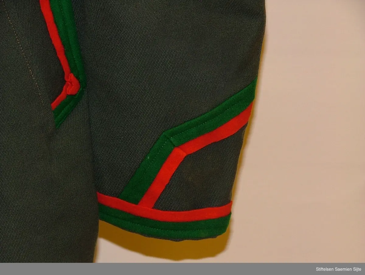 En dressjakke med slag og lommer som er blitt pyntet med røde og grønne kledesbånd. Foret med brunt bomullsstoff. Syv hekter i åpningen fram. Innerlomme