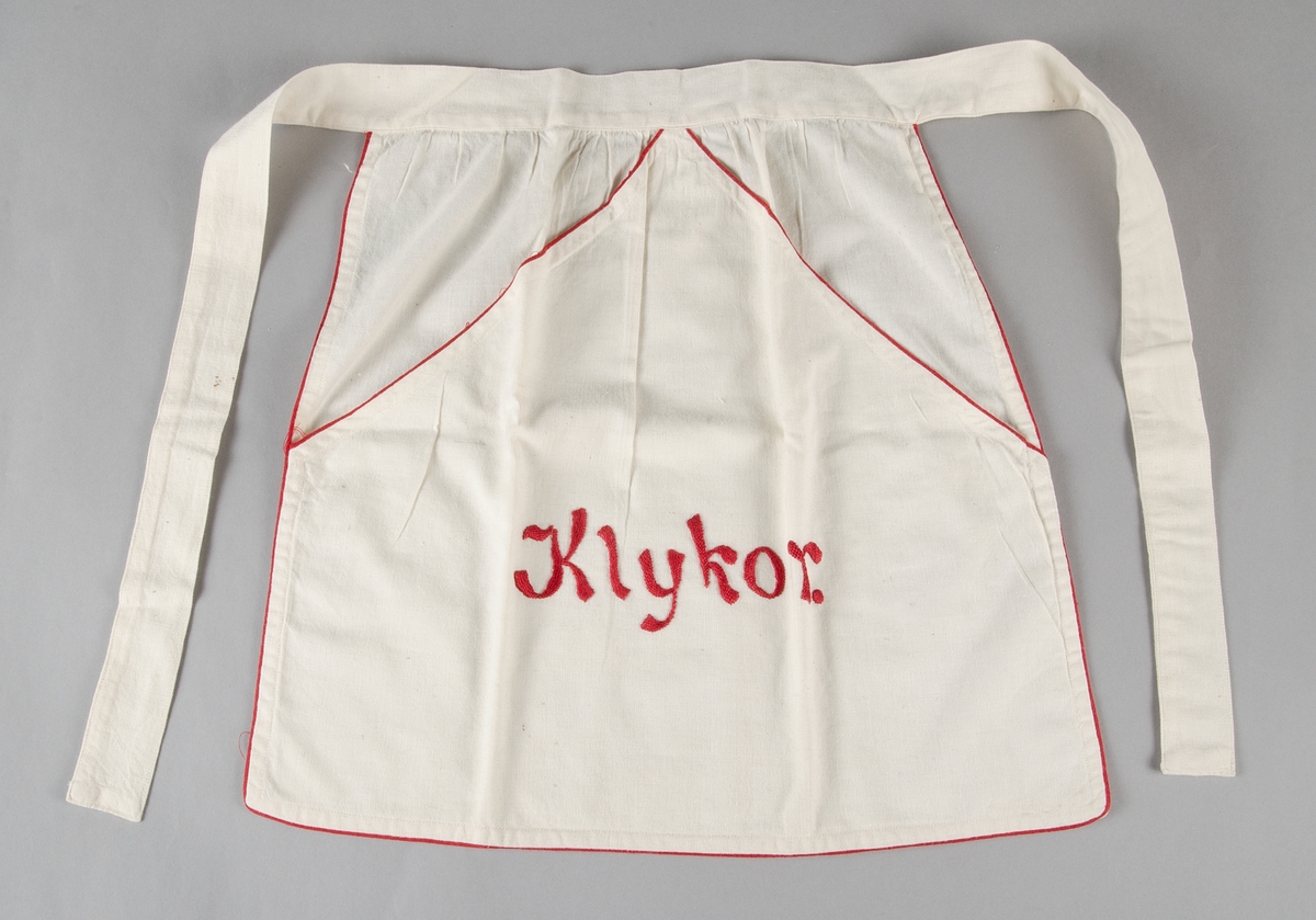 Kort midjeförkläde av vit bomullslärft med röd smal bandkant, med stor ficka med rött plattsömsbroderi: "Klyko". Linning och knytband i samma tyg.