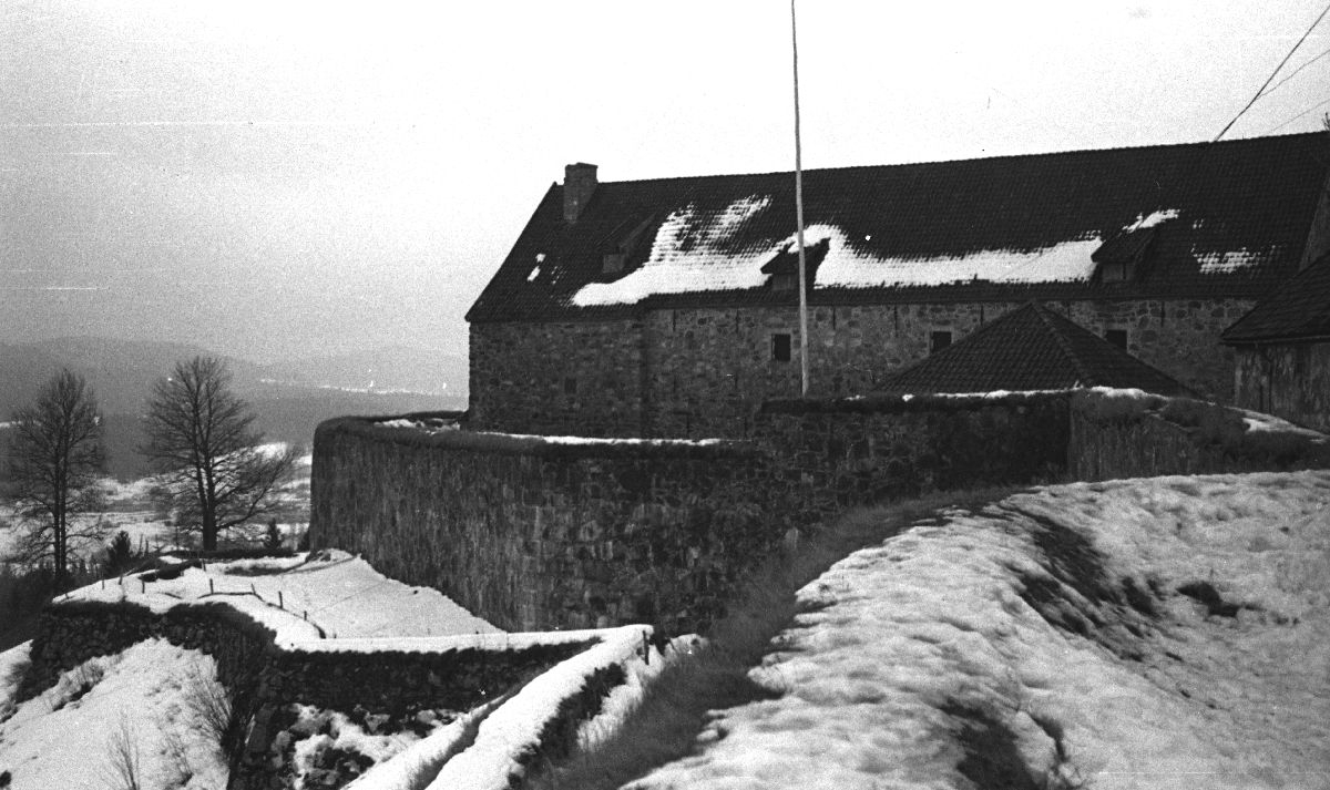 Landskap. En gammel steinbygning med en naken flaggstang beliggende på en høyde og med god oversikt over området. På bakken ligger snøen hvit.