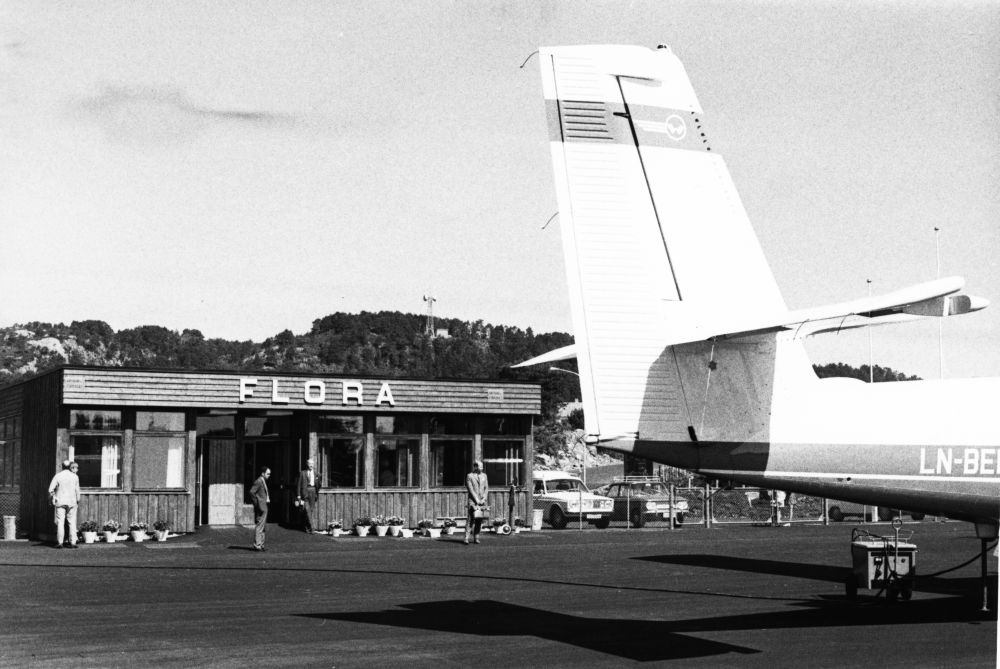 Lufthavn/Flyplass. Flora. Et fly, DHC-6 Twin Otter fra Widerøe, parkert foran flyekspedisjonen. Startaggregatet tilkoblet. Noen personer venter utenfor bygningen.