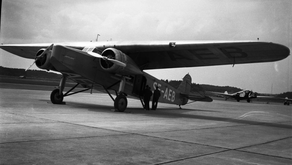 Lufthavn/Flyplass.  Et fly, SE-AEB, Fokker, parkert på oppstillingsplass. Tre personer samlet ved inngangsdøra. I bakgrunn et fly av typen Junkers Ju52.