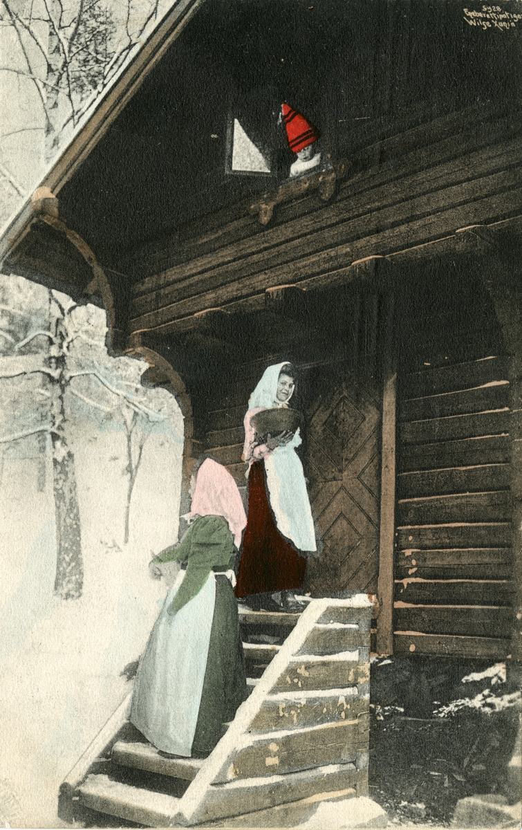 Kolorert postkort. To kvinner på trappa foran stabburet. Liten gutt med nisseluse titter ut fra vinduet.