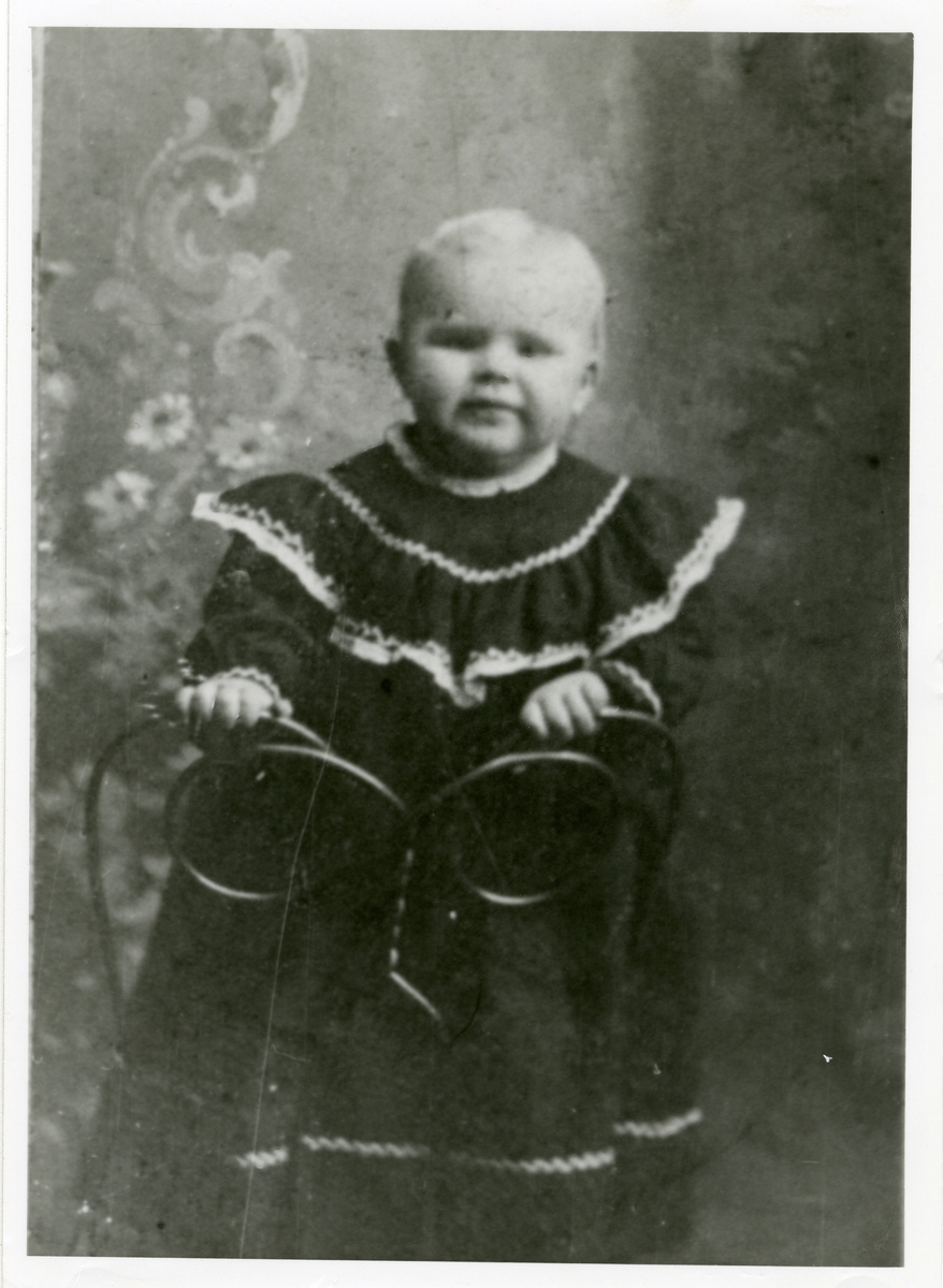 Portrett av en liten gutt. Gutten er iført kjole og står på en stol.
