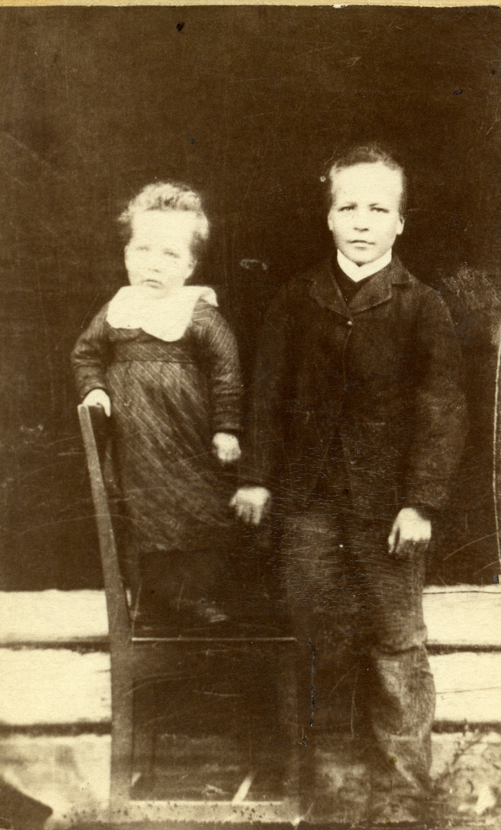 Portrett av en jente og en gutt. Jenta står på en stol og hun er iført en kjole. Gutten er iført jakke og bukse.