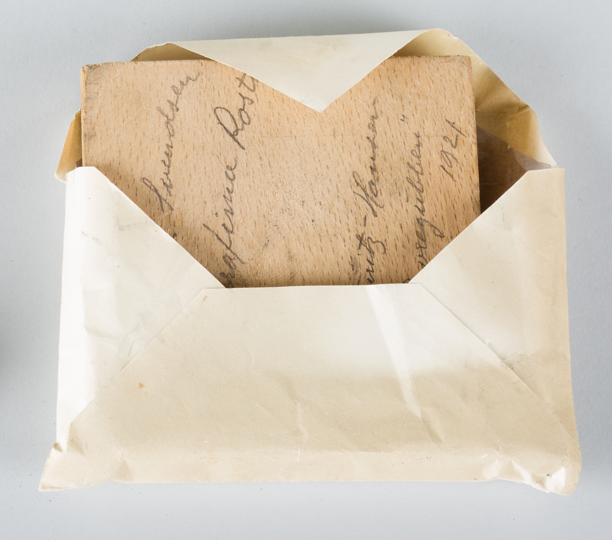 En kvadratisk trykkblokk av tre og metall, oppbevart i konvolutt av papir. 
Klisjéen er gitt i gave, en liten hilsen er påskrevet konvolutten.