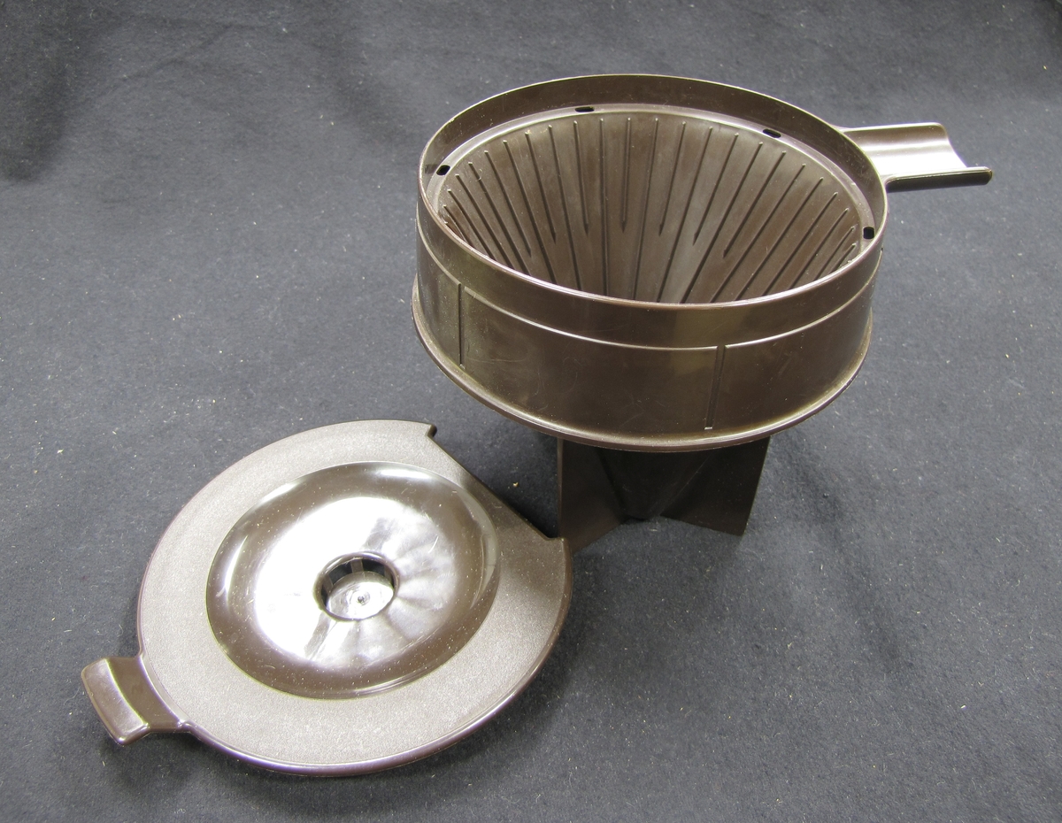 En av två stycken hållare för melitta - filter till kaffebryggning.

Bryggtratten är av brun plast och har ett lock.

Föremålen ingår i en samling från 1950 och 1960 - talet. som använts vid olika skolkök i Trollhättans kommun. Föremålen utrangerades på 1980 - talet och skänktes till Länsmuseet.