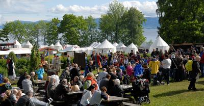 Folkehav på Hamar Middelalderfestival, med toppen av mange hvite middelaldertelt stikkende opp bak menneskene og Mjøsa helt i bakgrunnen.