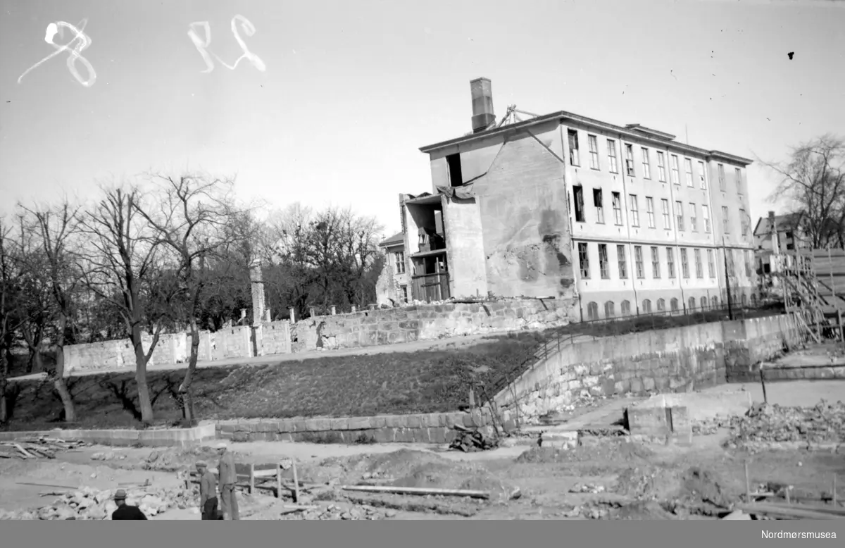 På bildet ser vi ruinene etter tyskernes bombing av Den Høgre Skoles trebygning i Kristiansund (mot tomta der Folkets Hus ble reist seinere). Den var bygget i 1870. På loftet i denne fantes bymuseet. Murbygningen til høyre på bildet, er fra 1923. Bildet er datert 14. juli 1940. Nordmøre Museums bildesamling


