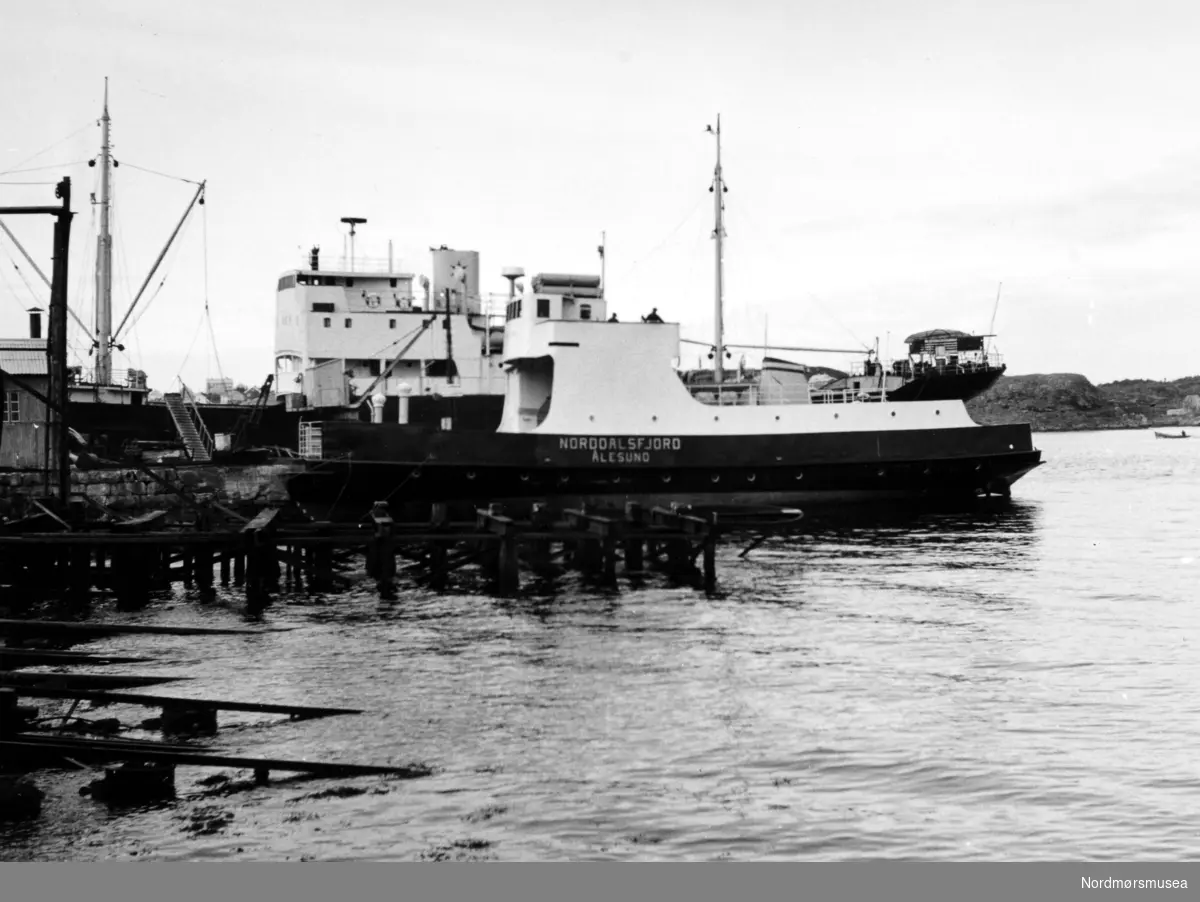 Bildet viser B/F";Norddalsfjord"; Storviks Mek. Verksted bnr. 14 ferdig utrustet ved piren. Båten vi ser bak ved piren er Ole T. Flakkes lastebåt M/S";Knoll";. "Norddalsfjord" ble levert til Møre og Romsdal Fylkesbåtar 15. mars 1961 og hadde følgende hoveddimensjoner: L 31,20 m x B 8,55 m x D 3,35 m og hadde en tonnasje på 159 brutto-registertonn. Fremdriftsmaskineriet består av 3 Volvo Penta turboladede dieselmotorer type TMD96 på til sammen 420 hk som via kilremdrift var koblet til et felles gir og propellaksel med vribar propell, slik at hver enkelt av motorene kunne kjøres separat. Fergen hadde 2 Bolinders vekselstrømsaggregater type 1052MG på 23 hk hver tilkoblet en generator på 17 kW. Fergen var utstyrt med elektrohydraulisk styremaskin. Fergen har plass til 18 personbiler og har sertifikat for 160 passasjerer. Forut er det innredet 6 lugarer for offiserer og restaurantpersonale og akterut en mannskapslugar for 4 personer og toppfarten er 11,4 knop og marsjfarten 10,5 knop. Ferga er verkstedets første nybygg etter B/F"Trygge" som ble levert i 1938. I forgrunnen ses bakenden på den nye klinkbygde patentslippvogna i stål fra 1918, den største slippen mellom Bergen og Trondheim, som ble erstattet med nytt slippanlegg ca. 1979, og banene til såpehellingen fra 1901 nærmest. Bygget helt til venstre inneholder en dampkjel og nederst i høyre hjørne ses litt av trekaia nedenfor støperiet. Bak ferga ses Teistholmen på Gomalandet. Bildet er fra 1961. Tekst av Peter Storvik. Fra Nordmøre museums fotosamlinger.



