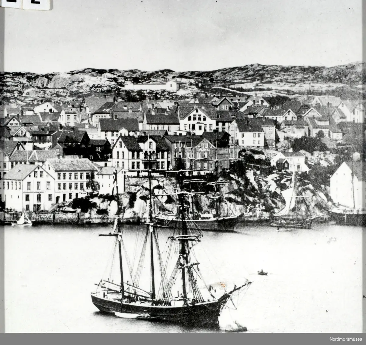 Bildet viser sansynligvis S/S " Ina" en skonnertbrigg på 252 registertonn fortøyd i "Storbøya" på havna i Kristiansund i 1885.
Bildet gir oss en fin oversikt om hvordan havnepartiet fra Kirkeallmenningen til omtrent der hvor Nordmørskafeen er i dag, var på den tiden. Vi ser at skutene ligger akterfortøyd ved Blixhammeren. Navnet på barken som ligger der er ikke kjent.
"Ina"s redere var N. R. Parelius og Albert Heyerdahl og fører var O. Tomelthy.
Skipet ble bygd i Genova i 1874.
Kildene forteller at hun var rundgattet og ikke videre pen, men sterk da den var bygd for kisfart. Seilte mest på Amerika og spesielt på Brasil. På Brasilkysten kom den ut for en kastevind som tok hele dokken, riggen og storstangen. Mannskapet fikk rigget nødrigg, og etter to uker greide de å komme seg tilbake til avseilingshavnen, som var Bahia. 
Her ble de liggende i fire måneder for reparasjon. Deretter gikk turen til London, hvor hun ankom den 15/7-1886. På den påfølgende turen over Atlanteren, under meget vanskelige og farlige forhold, berget de 10 mann fra en bark fra Rostock. Redningen ble utført av styrmann Alnæs sammen med tre matroser. Barken sank tre timer etter at man hadde fått de skipbrudne ombord i S/S "Ina". Skuta seilte inn til Falmouth med de overlevende. 
Korte glimt av "Ina"s seilingsliste viser at hun ankom Arkhangelsk den 24/7-1885 og gikk derfra den 12/8 til Davenport. Fra Trinidad gikk hun den 8/2-1887 til Falmouth. Kapteinen var O. Tomelthy.
Det fremgår, som kildene sier, at hun var noe rundgattet, men man har vanskelig med å si seg enig i at hun ikke var pen.
S/S " Ina" ble i 1887 solgt til Porsgrunn.                                                                                 Kilde : Kristiansund på de 7 hav av Kjell Dønheim.                                                                   Se FAKf-100296.187028    (KMb-1987-005.3763)                                                                    Se FAKf-100296.187121    (KMb-1987-005.3854)                                                                        Se FAKf-100296.187127    (KMb-1987-005.3860)                                                                     Se FAKf-100296.198996    (KMb-1987-005.0428)                                                                                                                                                            Se FAKf-100296.201075    (KMb- 1992-030.0020)                                                                     Se FAKf-100296.214272    (KMb-1946-000.0859)                                                                                 Se FAKf-100296.220923    (KMb-1987-005.5884)
(Info: Peter Storvik). -- Foto fra havnebassenget i Kristiansund og bebyggelsen på Kirklandet. Bakerst i bildet sees barkskipet "Ina" fortøyd ved Blixhammeren. Skipet i front er ikke kjent.  Bildet er trolig fra perioden 1880 til 1900. Fotograf er ukjent. Fra Nordmøre Museums fotosamlinger.