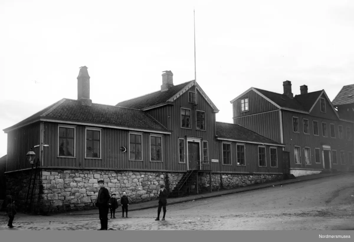 Bernstorffskolens "Sømandsskole". I 1905 ble det vedtatt å selge Bernstorffskolen med 1/2 mål grunn til Norges Bank for 15.000 kroner. Til da hadde Norges Bank holdt til i Sparebank-gården i Skolegaten, som var byens "finansgate" på den tiden. De andre bankene hadde også kontor i denne gaten. Bygningen ble tegnet av Kristen Tobiaz Rivertz fra Kristiania, som oppholdte seg da i perioden 1907 til 1909 i Kristiansund, hvor han blandt annet har tegnet "en Badeanstalt", Festiviteten, Grand Hotell og Norges banks lokaler. Norges Banks nye lokaler stod ferdig i 1909, hvor de stod frem til 28. april 1940 da banken brant ned sammen med store deler av byens bebyggelse. Da krigen var over ble det nye Norges Bank oppført i perioden i 1945-1952 og ble da tegnet av Arkitektene Eivind Moestue og Ole Lind Schistad. I dag (2007) holder Kristiansund folkebibliotek til i dette bygget. Ellers så ser vi Folkets hus til høyre for Bernstorffskolen. I 1915 flyttet Tidens Krav inn her i samme lokaler med sitt trykkeri. Her holdt de til inntil økonomien ble for trang, og i 1924 flyttet de ut av lokalene og inn i nabobygget - igjen til høyre - Arbeiderforeningen. 
Fra Nordmøre Museums fotosamlinger.
Kilde (Bl.a): Eivind Aass: Kristiansunds Historie VI, side 43
Kilde: Petter E. Innvik: Et fyrtårn i havgapet. Tidens Krav gjennom 100 år. 1906 - 2006, side 41 og 47.