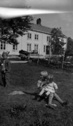 Tre barn på en plen foran et gårdshus. En bil er parkert i b