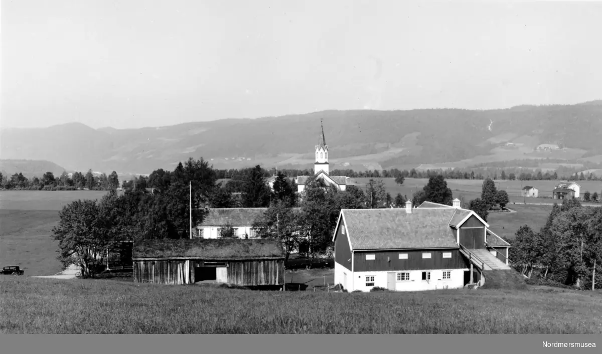 Foto fra Rindal prestegård i Rindal kommune. Her ser vi gården i front med Rindal kirke i bakgrunnen. Fotograf og datering er trolig Georg Sverdrup og datering er sannsynligvis fra perioden 1930 til 1939. Fra Nordmøre Museums fotosamlinger. Reg: EFR
