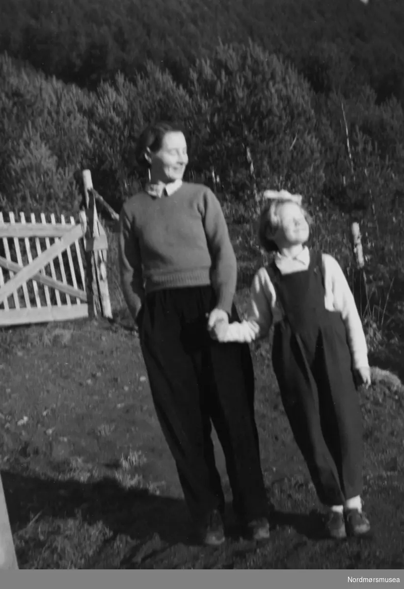 Her ser vi en kvinne sammen med en pike ved en boligport. Trolig enten familie, slekt eller bekjente av fotografen, som sannsynligvis er John Myren. Datering er ukjent, men trolig mellom 1930 til 1960. Stedet er også ukjent, men kan være fra Sunndal kommune. Fra Nordmøre museums fotosamlinger. 
