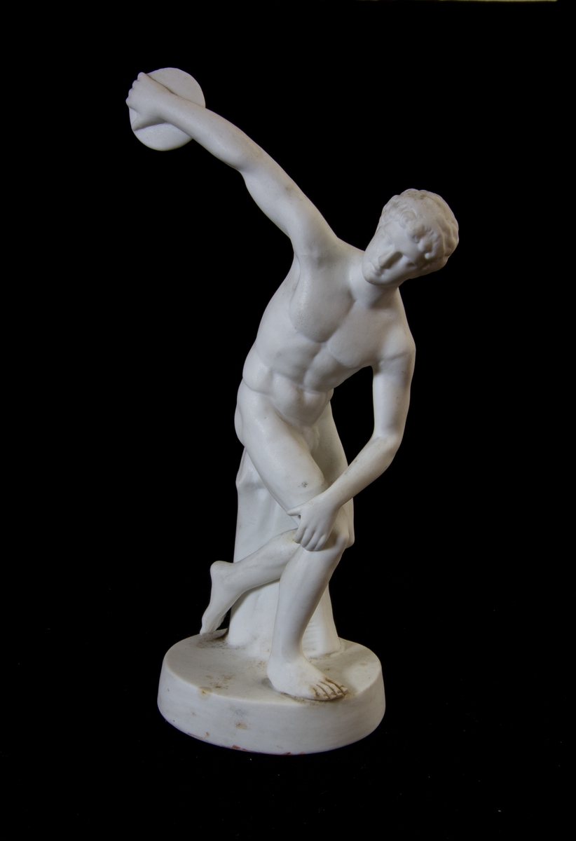 Studiemodell i parian föreställande en naken man i helfigur som håller en diskus i sin bakåtsträckta hand. Lutar sig delvis mot ett stubbliknande föremål. Oval fotplatta.