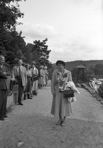 En kvinna står på en trädgårdsgång. Till vänster syns flera människor. I bakgrunden skymtar en trädgård med trädgårdsmöbler.