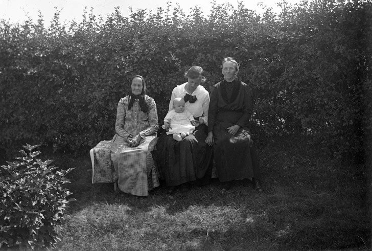 Några kvinnor sitter på en trädgårdsbänk, en av dem med ett barn i knäet, intill en häck. 
Troligen fr. v.: fru Christina Svensdotter (1831-1923) (mormor t. Fritz Bergqvist), Agda Bergqvist med sonen Valter, fru Carolina Sofia Bergqvist (1861-1939) (mor till Fritz B.).