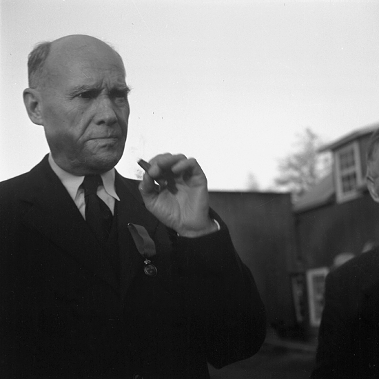 Närbild av en äldre man med mörk kostym, som röker en cigarett ...
